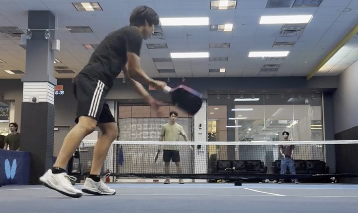 A frame of real doubles play: the near player mid-swing with the paddle on the ball, the net and the far side of the court behind them.